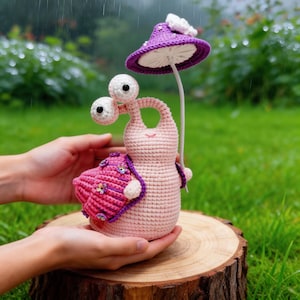 Puede incluir: Figura de caracol de ganchillo en tonos rosa, morado y blanco. El caracol tiene ojos grandes y saltones y un paraguas con forma de seta. La figura se sostiene sobre una superficie de madera, con un fondo verde.