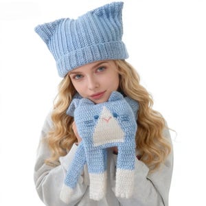 May include: A light blue knitted cat ear hat and matching scarf. The scarf features a cat face with white and blue details, including a pink mouth and blue eyes. The hat has a ribbed design.