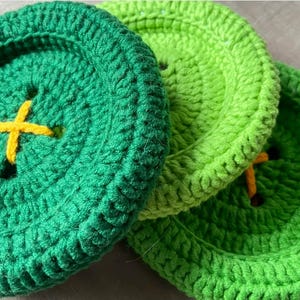 May include: Three circular crochet coasters designed to resemble large buttons. The coasters are made in shades of dark green and light green yarn, each featuring a central yellow cross-stitched detail that mimics the thread of a button.