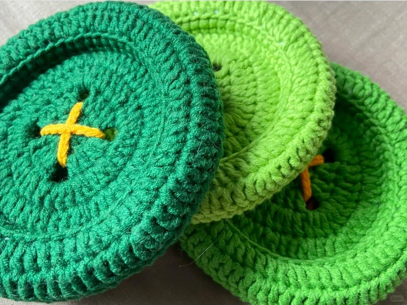 May include: Three circular crochet coasters designed to resemble large buttons. The coasters are made in shades of dark green and light green yarn, each featuring a central yellow cross-stitched detail that mimics the thread of a button.