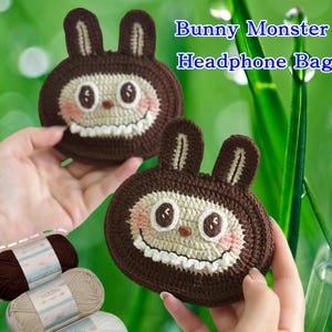 Puede incluir: Dos bolsas de ganchillo marrones para auriculares con un diseño de monstruo conejito se sostienen en alto. Las bolsas tienen una cara blanca, mejillas rosas y una boca sonriente. El texto "Bunny Monster Headphone Bag" está en el fondo. Hilo en la esquina inferior izquierda.