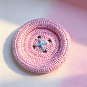 May include: A round, crocheted button in a soft pastel pink yarn. The button features a raised outer rim and a recessed center with four holes arranged in a square pattern. A light blue yarn is stitched through the holes in an X shape.