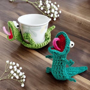 May include: Two handmade crochet lizard figures in vibrant green yarn. One lizard is wrapped around a white ceramic cup with a palm leaf pattern, while the other stands upright. Both feature wide-open red mouths, pink tongues, and white eyes.