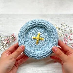 May include: A round, light blue crocheted object shaped like a large button, featuring a textured surface and a central yellow cross-stitched detail made of yarn, creating a circular design with a raised border and a recessed center.