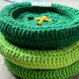 May include: A stack of three circular, crocheted coasters in varying shades of green. The top coaster is dark green with a small, yellow, cross-stitched detail in the center and a small white fabric tag attached to the outer edge.