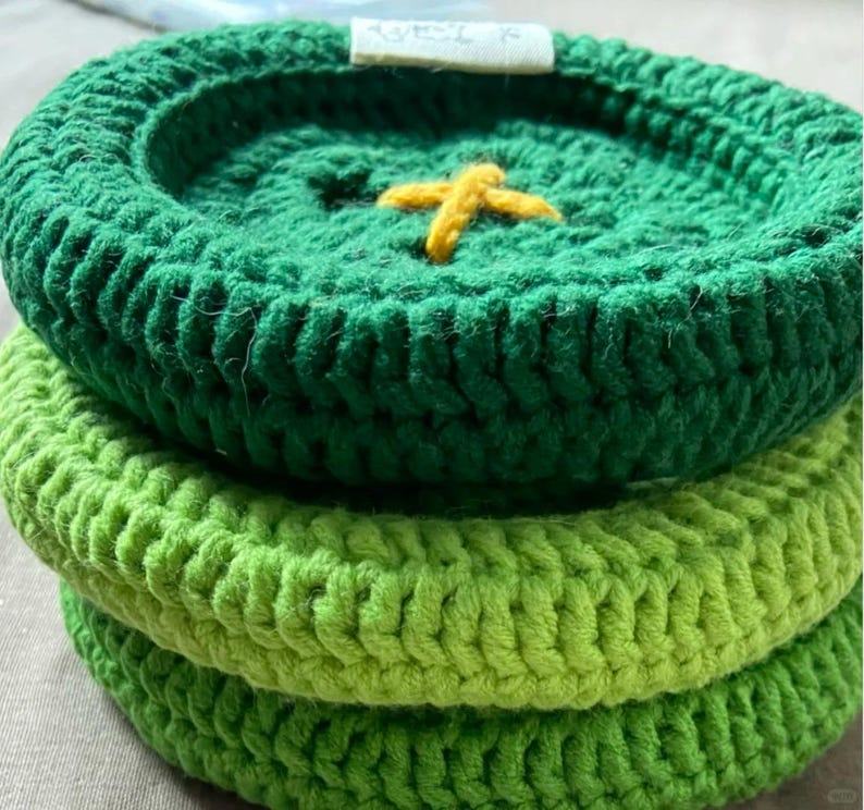 May include: A stack of three circular, crocheted coasters in varying shades of green. The top coaster is dark green with a small, yellow, cross-stitched detail in the center and a small white fabric tag attached to the outer edge.