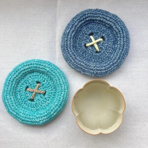 May include: Two circular crocheted coasters shaped like large buttons, one in medium blue with a pale yellow cross-stitch center and one in turquoise with a tan cross-stitch center, alongside a small, cream-colored, flower-shaped ceramic bowl.