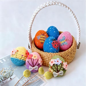 May include: A collection of colorful, crocheted Easter eggs, some with floral and insect designs, are displayed in a woven basket with a white handle. Additional eggs with floral embellishments are arranged nearby, alongside decorative yellow pom-poms.