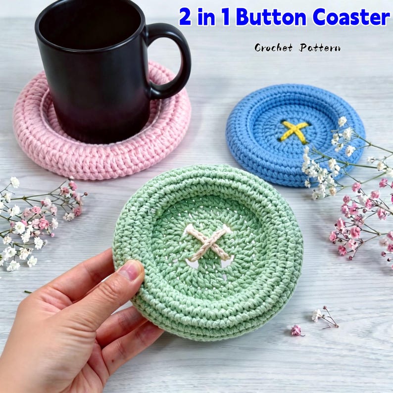 May include: Three round crochet coasters shaped like buttons in light pink, light blue, and light green. The pink coaster supports a black mug. The blue and green coasters feature central cross-stitch detailing. Text reads 2 in 1 Button Coaster Crochet Pattern.