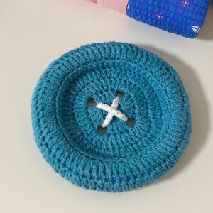 May include: A large, circular, teal-colored crochet button featuring a recessed center with four holes stitched with white yarn in an X pattern. The button has a thick, textured, hand-stitched appearance with a prominent raised outer rim.