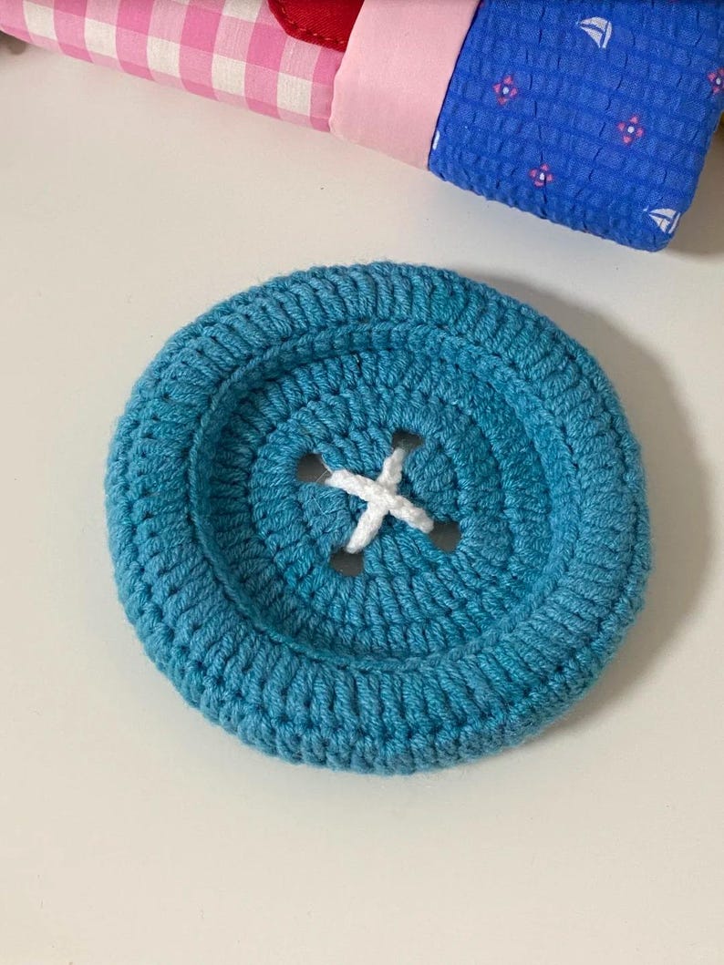 May include: A large, circular, teal-colored crochet button featuring a recessed center with four holes stitched with white yarn in an X pattern. The button has a thick, textured, hand-stitched appearance with a prominent raised outer rim.