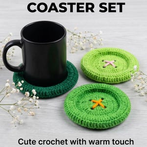 May include: A coaster set featuring three round, button-shaped crochet coasters in shades of dark green and light green. One dark green coaster supports a black ceramic mug. The light green coasters have pink and orange yarn cross-stitch details.