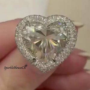 Puede incluir: Un anillo de diamantes en forma de corazón con un halo de diamantes más pequeños. La piedra central está facetada, reflejando la luz. El anillo se sostiene, mostrando su brillo. El texto "SparkleHouseCA" es visible.