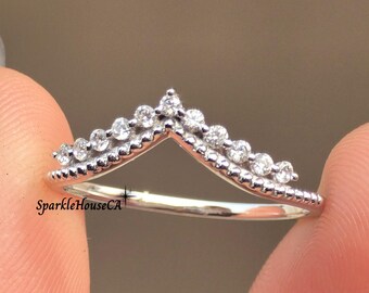 May include: A silver-colored ring in a V-shape, adorned with small, clear gemstones. The ring's design resembles a crown or tiara. The text "SparkleHouseCA" is visible on the ring. The ring is held between two fingers.