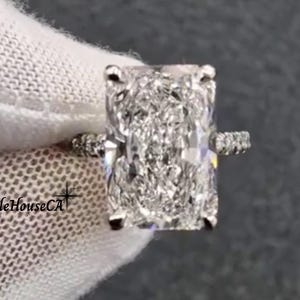 May include: A radiant-cut diamond ring with a pavé band. The large, rectangular-shaped diamond is the focal point, set in a classic prong setting. The band is adorned with smaller diamonds. The ring is held against a white fabric background.