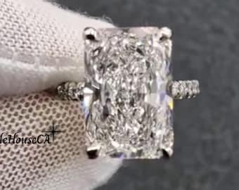 May include: A radiant-cut diamond ring with a pavé band. The large, rectangular-shaped diamond is the focal point, set in a classic prong setting. The band is adorned with smaller diamonds. The ring is held against a white fabric background.