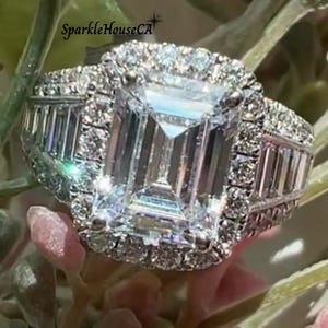 May include: A sparkling diamond ring featuring an emerald-cut central stone, encircled by a halo of smaller diamonds and baguette-cut stones on the band. The ring is displayed amidst green and pink foliage, creating a luxurious and elegant aesthetic. The text "SparkleHouseCA" is visible.