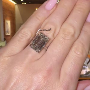 May include: A large, rectangular-cut diamond ring. The ring features a clear, emerald-cut diamond set on a thin, silver band. The ring is on a hand with pink nail polish.