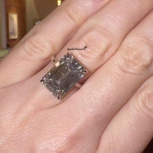 May include: A large, emerald-cut diamond ring with a silver band. The diamond is a light gray color and is set with small accent stones. The ring is on a finger. The text "SparkleHouseCA" is visible.