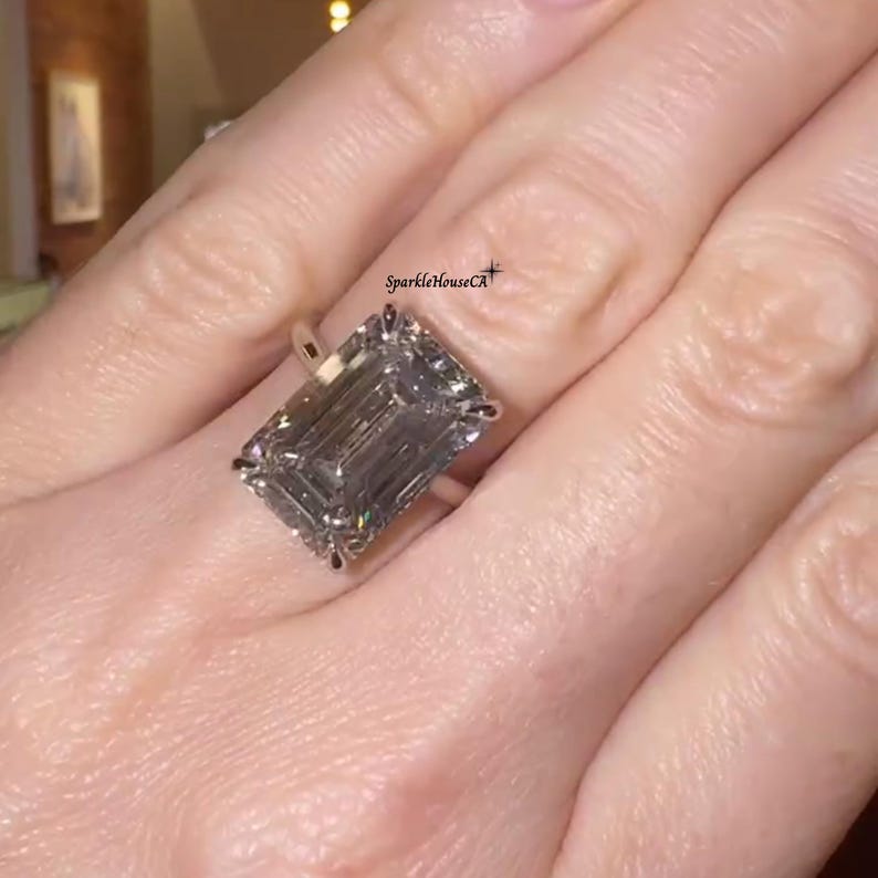 May include: A large, emerald-cut diamond ring with a silver band. The diamond is a light gray color and is set with small accent stones. The ring is on a finger. The text "SparkleHouseCA" is visible.