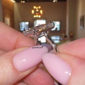 May include: A close-up of a silver ring with a large, rectangular, clear gemstone. The ring is held between two fingers with pink nail polish. The ring's band is thin and elegant, and the gemstone is set with smaller stones.
