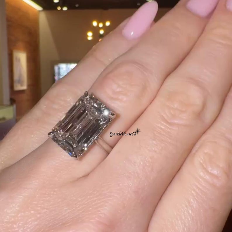 May include: A large, rectangular-cut diamond ring. The diamond is set on a thin, silver band. The ring is shown on a person's finger. The background is blurred, but appears to be indoors. The text "SparkleHouseCA" is visible.