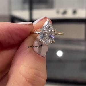 May include: A gold band engagement ring featuring a large, pear-cut diamond. The diamond is clear and sparkling, held in place by small prongs. The ring is presented to highlight its elegant design.