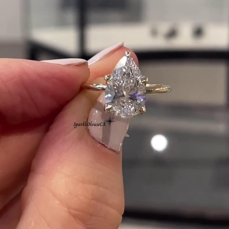 May include: A gold band engagement ring featuring a large, pear-cut diamond. The diamond is clear and sparkling, held in place by small prongs. The ring is presented to highlight its elegant design.