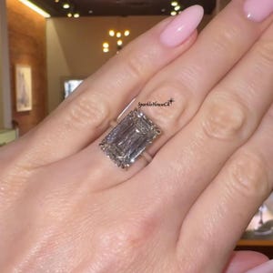 May include: A large, rectangular-cut diamond ring with a silver band. The diamond is clear and reflects light. The ring is on a hand with pink nail polish. The text "SparkleHouseCA" is visible.