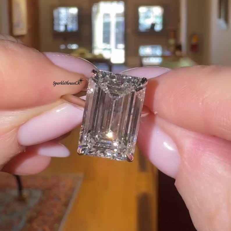 May include: A large, rectangular-cut diamond ring with a gold band. The diamond is clear and faceted, reflecting light. The ring is held up close, showcasing the sparkle and clarity of the gemstone. The text "SparkleHouseCA" is visible.