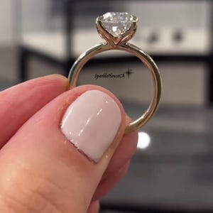 May include: A gold ring featuring a large, clear, round-cut gemstone. The ring is held up, highlighting the gemstone's facets and the slender gold band. The ring is a piece of jewellery.