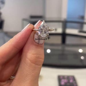 May include: A gold ring with a large, pear-cut diamond. The diamond is clear and sparkling. The ring is held up by a hand with manicured nails. The text "SparkleHouseCA" is visible on the image.
