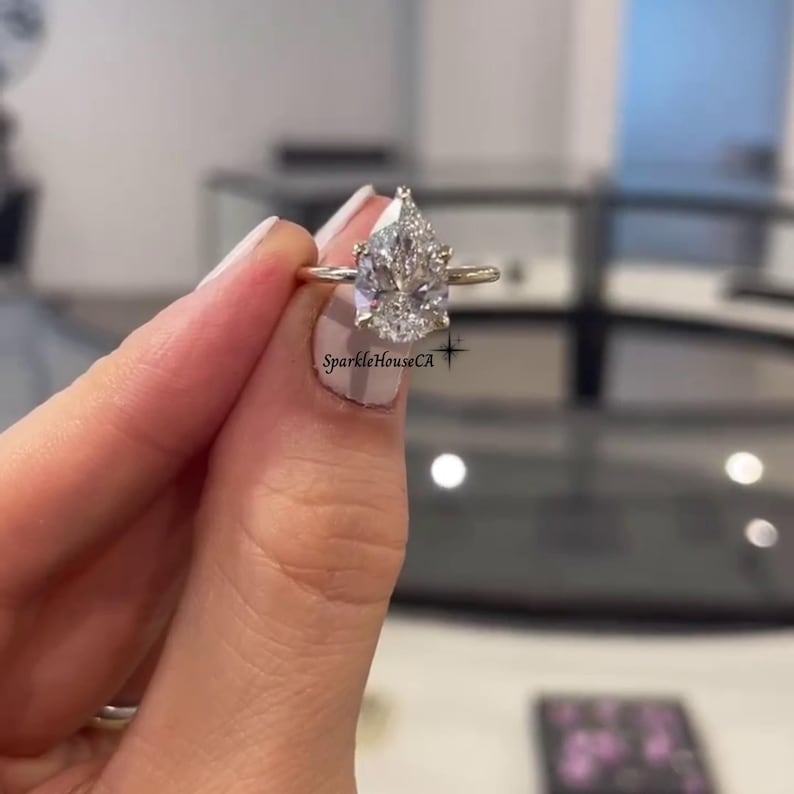 May include: A gold ring with a large, pear-cut diamond. The diamond is clear and sparkling. The ring is held up by a hand with manicured nails. The text "SparkleHouseCA" is visible on the image.