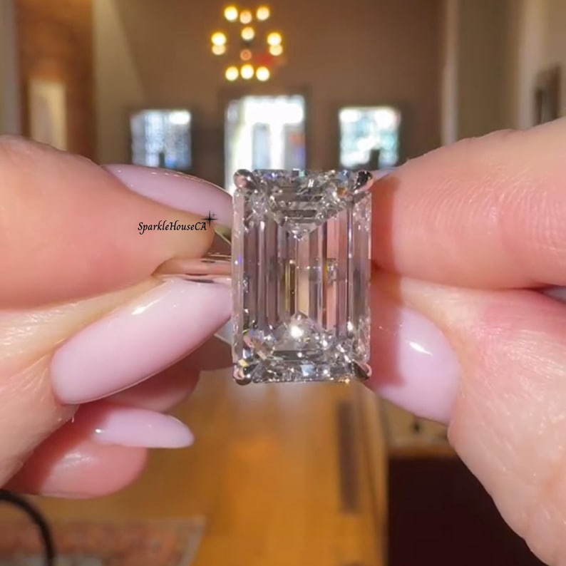 May include: A large, rectangular-cut diamond ring. The diamond is held between two hands with pink nail polish. The ring is highly reflective, with visible facets. The text "SparkleHouseCA" is visible on the ring.