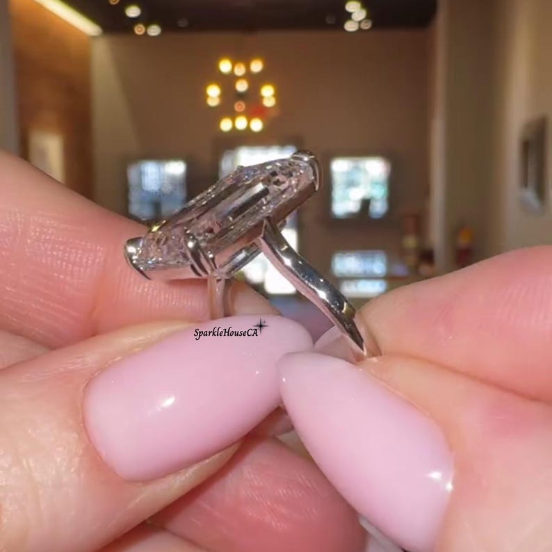 May include: A close-up of a silver ring with a large, rectangular, clear gemstone. The ring is held between two fingers with pink nail polish. The text "SparkleHouseCA" is visible on the ring.