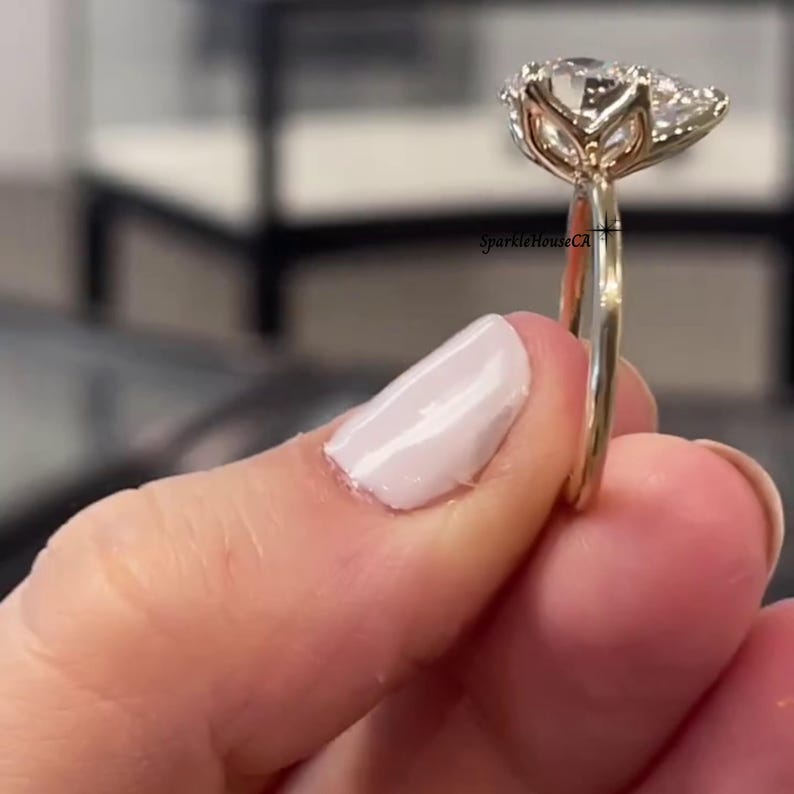 May include: A gold engagement ring with a large oval-cut diamond centre stone, flanked by smaller accent stones. The ring's band is plain, polished gold. The ring is held up, highlighting its design and sparkle.