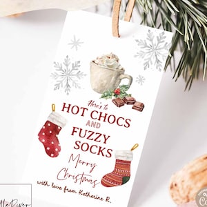 May include: A white gift tag with a watercolor illustration of a mug of hot chocolate, two Christmas stockings, and snowflakes. The tag reads "Here's to Hot Chocs and Fuzzy Socks Merry Christmas". The tag is tied with a brown string.