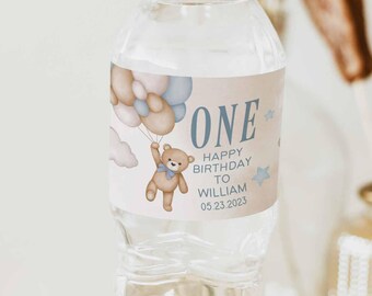 May include: A water bottle with a custom label for a first birthday. The label features a teddy bear holding balloons, the word "ONE", and the text "HAPPY BIRTHDAY TO WILLIAM 05.23.2023". The label has a light blue and beige color scheme.