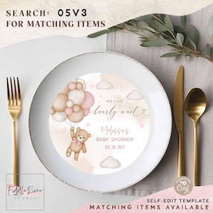 May include: A white plate with a baby shower design. The plate features a teddy bear holding balloons, with text that reads "We can bearly wait Melissa's Baby Shower Dec 20, 2022". Gold cutlery is placed to the side of the plate.