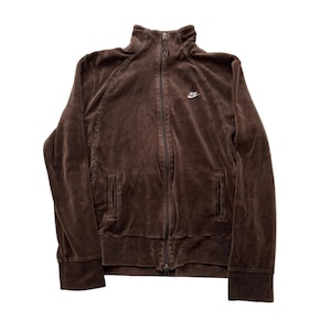 May include: A brown zip-up jacket with a small white Nike logo on the chest. The jacket has a high collar, two front pockets, and ribbed cuffs and hem. The jacket is made of a soft, velvety material.