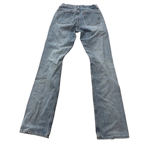 Könnte beinhalten: Hellblaue Jeans mit klassischem Fünf-Taschen-Design. Die Jeans haben einen geraden Beinverlauf und werden von hinten betrachtet. Der Denim hat eine verwaschene Optik. Die Jeans liegen flach auf einer weißen Oberfläche.