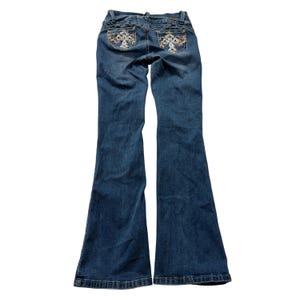 Op de afbeelding: Blauwe spijkerbroek van denim met uitlopende pijpen en een taille met gemiddelde hoogte. De achterzakken zijn versierd met ingewikkelde wit en oranje geborduurde kruisontwerpen, geaccentueerd met kleine doorzichtige strass-steentjes.