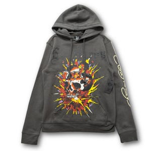 May include: A charcoal gray hooded sweatshirt featuring a large, vibrant graphic of a skull surrounded by yellow, orange, and red starburst patterns on the front. The sleeve displays cursive text, and a black brand tag hangs from the front pocket.