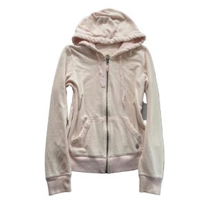 May include: A pale pink zip-up hoodie featuring a soft texture, a drawstring hood, and a full-length metal zipper closure. The garment includes two front pouch pockets and ribbed cuffs at the sleeves and hem, with a small metallic logo emblem.