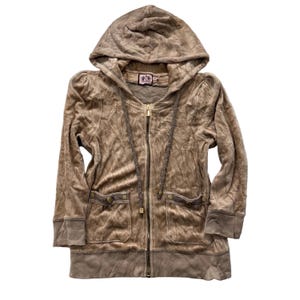 May include: A tan velour zip-up hoodie with a hood and pockets. The hoodie features a drawstring and a small emblem near the neckline. The zipper and pocket details are gold-toned. The fabric has a textured appearance.