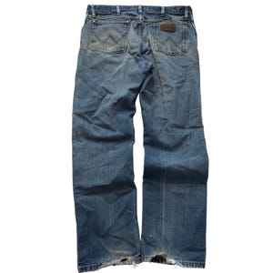 May include: Faded blue denim jeans with worn areas and distressed details. The jeans feature classic five-pocket styling, a brown leather patch, and the Wrangler logo. The back pockets have a signature W-shaped stitching.