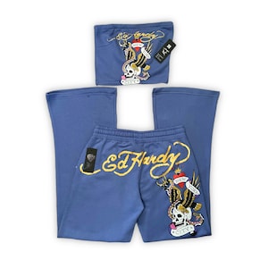 May include: A blue two-piece tracksuit set with the Ed Hardy logo in gold script. The top is a strapless tube top, and the trousers are flared. Both items have a graphic of a skull, eagle, and the words "New York City".