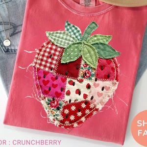 May include: A pink t-shirt featuring a patchwork strawberry design. The strawberry is crafted from diverse patterned fabrics, including gingham, floral, and heart prints. The shirt is labelled "Crunchberry" and is shown with jeans and a hat.