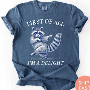 May include: Denim blue t-shirt with a white graphic of a raccoon dancing. The text "FIRST OF ALL" is above the raccoon, and "I'M A DELIGHT" is below. The shirt is a casual, unisex style.