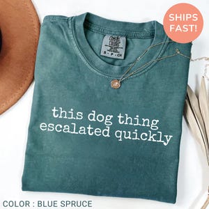 May include: A blue spruce colored t-shirt with white text that reads this dog thing escalated quickly. The shirt features a crew neckline and a Comfort Colors brand label on the inside collar. A thin gold chain necklace with a small round pendant rests on top.
